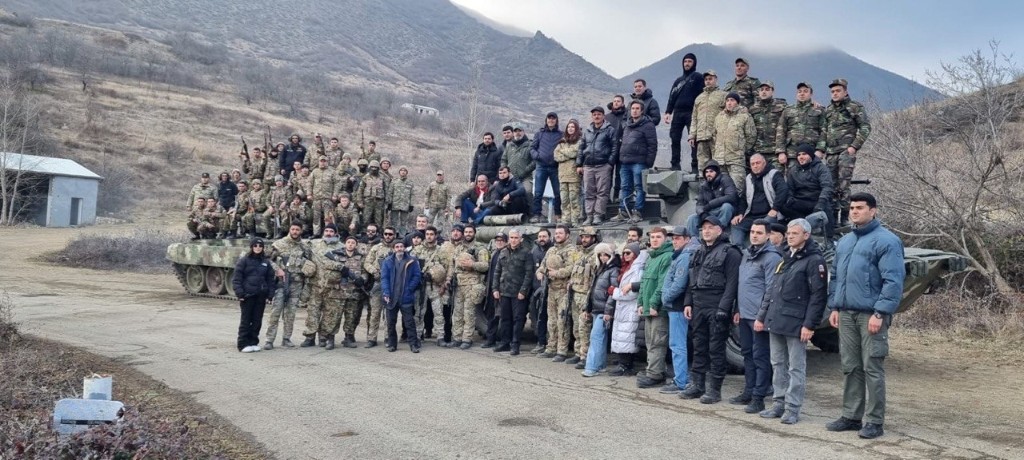44 cast and crew group photo around a camouflaged armored vehicle